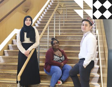 Three young students facing forward and smiling in front of City's indoor stairwell