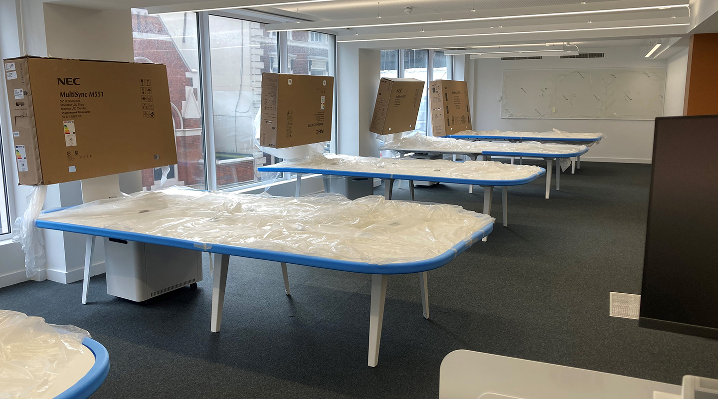 Learning space with group-working desks still covered in dustsheets. Monitors at the end of each desk still have their boxes over the top to protect them.