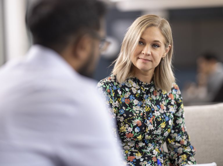 Two Bayes students, a man and a woman, having a conversation seated in the Bayes study area. The woman is in focus, while the man has his back to the camera and is out of focus, as is the background.