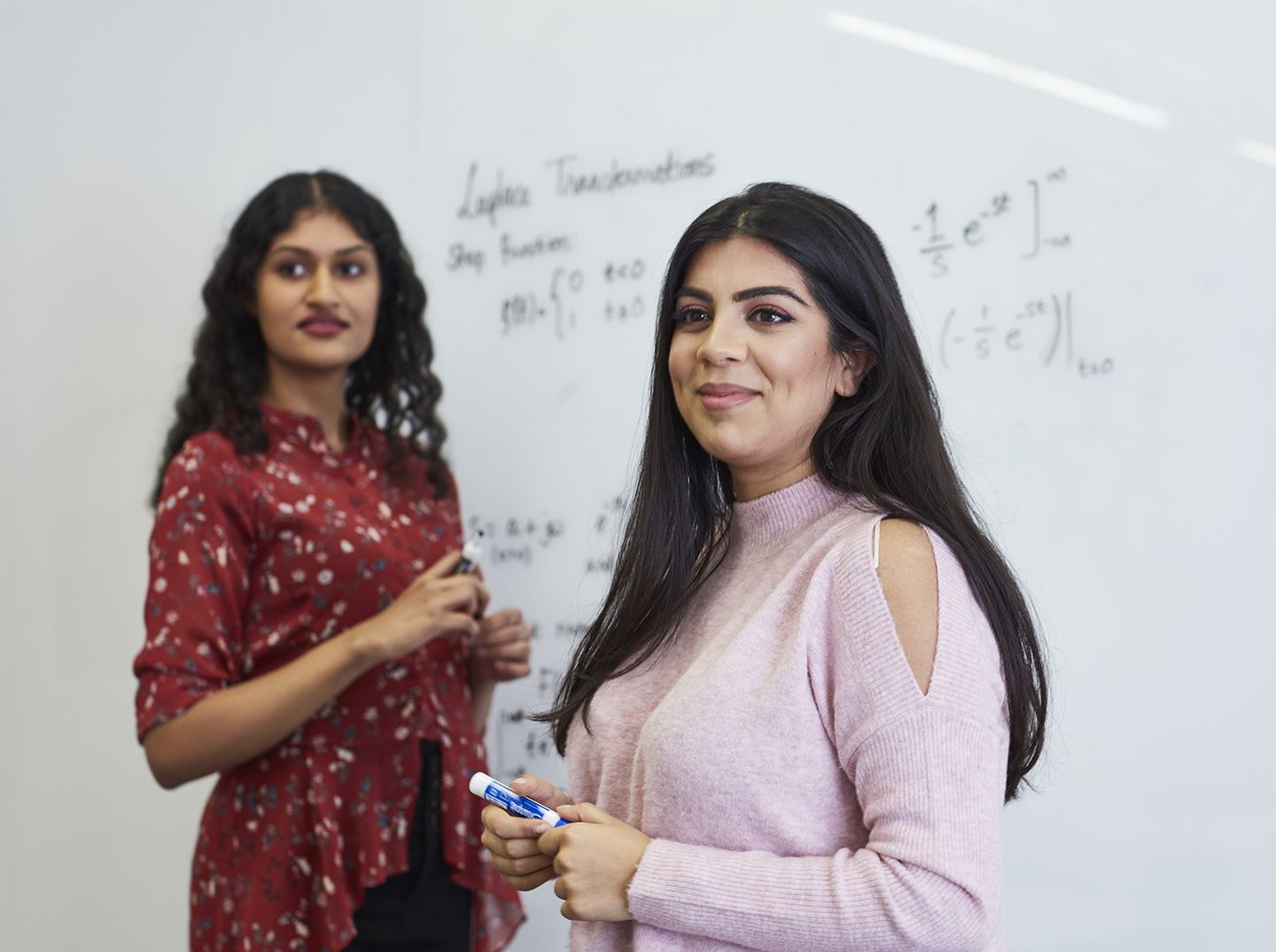 Two female students with whiteboard markers in front of a whiteboard with maths equations written on it.