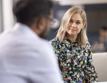 Two Bayes students, a man and a woman, having a conversation seated in the Bayes study area. The woman is in focus, while the man has his back to the camera and is out of focus, as is the background.