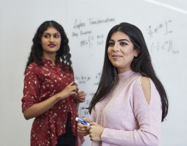 Two female students with whiteboard markers in front of a whiteboard with maths equations written on it.