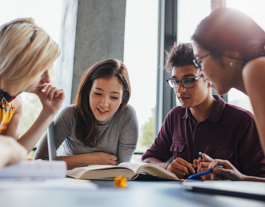 Four students around a table chatting and writing on paper. 